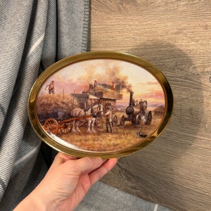 Puede incluir: Un plato ovalado de cerámica de estilo vintage con un borde dorado presenta una pintura de una máquina de vapor tirando de un vagón cargado de heno. La escena se desarrolla en un paisaje rural con una puesta de sol en el fondo.