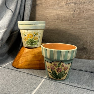 May include: Two painted terracotta flower pots with floral designs. The top pot has a blue and white glaze with yellow flowers. The bottom pot has a green and white glaze with purple flowers.