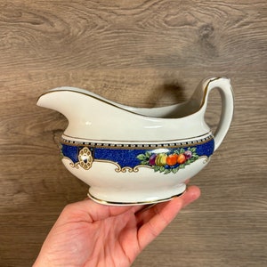 Puede incluir: Una salsera de porcelana blanca con un diseño floral azul y dorado. La salsera tiene una base ancha y redondeada y un pico en un lado. El asa está en el lado opuesto al pico.