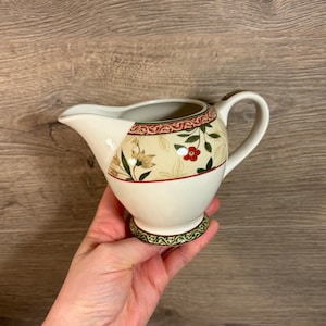 Può includere: Un creamer in ceramica bianca con un design floreale in tonalità di verde, rosso e marrone. Il creamer ha un manico e una base con un bordo verde e dorato.