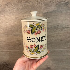 Może przedstawiać: Biały ceramiczny słoik na miód z kwiatowym wzorem i złotą obwódką. Słoik ma napis "HONEY" na przodzie.