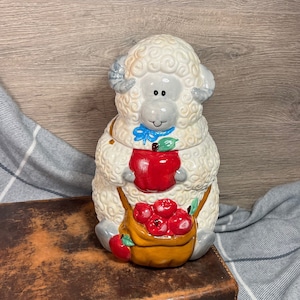 May include: A white ceramic sheep cookie jar with a blue bow and a basket of red apples. The sheep has a gray face and a friendly expression.