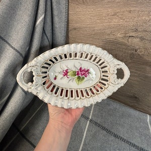 May include: White porcelain oval serving dish with a delicate openwork design and a floral motif in pink and green. The dish has two handles on the sides.
