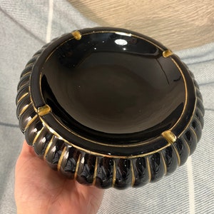 以下が含まれることがあります： 光沢のある黒い灰皿と金色のアクセント。 灰皿は側面にリブのデザインがあり、縁とタバコ置きに金色のディテールがあります。 灰皿は丸く、セラミック製のように見えます。