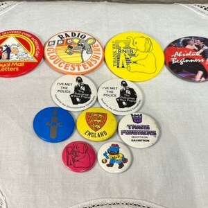Puede incluir: Una colección de diez botones vintage con varios diseños. Los botones presentan una variedad de colores, incluyendo rojo, azul, amarillo, naranja y verde. Algunos de los botones tienen texto, como "Royal Mail Letters", "Radio Gloucestershire", "RNIB", "I've Met The Police", "England", y "Transformers".