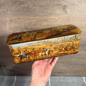 May include: Rectangular tin box with a lid featuring a hunting scene with riders on horseback, dogs, and a landscape. The box has a brown and gold color scheme, with the hunting scene depicted in various colors. The box is held by a hand.