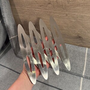 May include: A set of four silver-colored hair clips. Each clip has a long, slender shape with a rounded tip and a central oval cutout. The clips are held in a hand, with a gray and white striped fabric in the background.