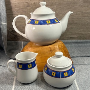 Puede incluir: Una tetera, una jarra de crema y un azucarero blancos con un patrón floral azul y amarillo. La tetera tiene una tapa y un pico. La jarra de crema tiene un asa. El azucarero tiene una tapa.