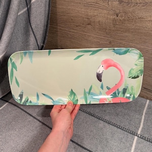 May include: Rectangular serving platter with a flamingo design. The platter features a light green background with a pink flamingo and green foliage. The edges are rounded, and the overall design is tropical.