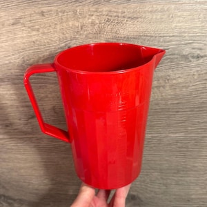 May include: A vibrant red plastic measuring jug with a handle and a pouring spout. The jug features measurement markings. The handle is rectangular and attached to the side of the jug.