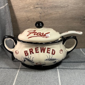 Puede incluir: Cafetera de cerámica blanca con tapa negra y asa negra. La cafetera tiene una inscripción roja que dice "Fresh Brewed" y una ilustración en blanco y negro de tazas de café.