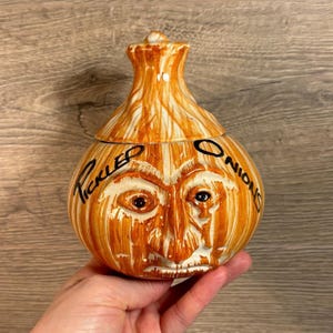 May include: A ceramic onion-shaped jar with a lid, painted in brown and white with a face and the words "PICKLED ONIONS".