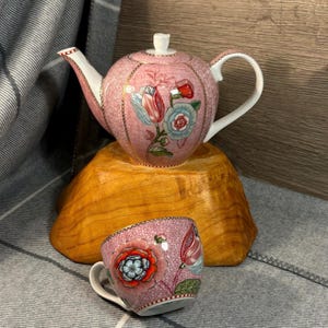 May include: A pink floral teapot and matching teacup with a delicate floral pattern. The teapot has a lid and a spout, and the teacup has a handle. Both pieces are sitting on a wooden surface.