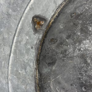 Op de afbeelding: Een close-up van een gegalvaniseerd metalen oppervlak met een cirkelvormige ring in reli&euml;f en een duidelijke vlek van oranjebruine roest. De textuur van het metaal is grijs en gevlekt, met tekenen van verwering en oxidatie.