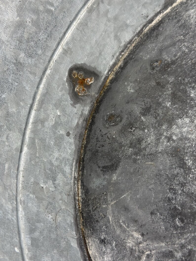 Op de afbeelding: Een close-up van een gegalvaniseerd metalen oppervlak met een cirkelvormige ring in reli&euml;f en een duidelijke vlek van oranjebruine roest. De textuur van het metaal is grijs en gevlekt, met tekenen van verwering en oxidatie.