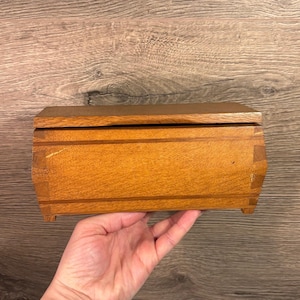 May include: A small, rectangular wooden box with a hinged lid. The box is made of light brown wood with visible grain and is held in a hand. The box has a curved top and is resting on small feet.
