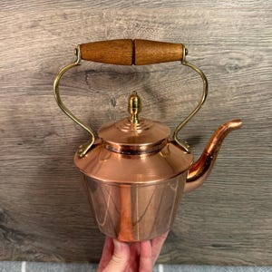 Op de afbeelding: Een koperen theepot met een houten handvat en messing accenten. De ketel heeft een afgeronde body, een tuit en een deksel met een kleine knop. De handgreep is gemaakt van twee stukken hout.