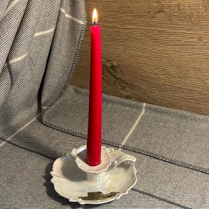 May include: A white ceramic candle holder with a handle and a red candle burning inside. The candle holder has a floral design and a small image of a building on the bottom.