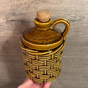 Puede incluir: Jarra de cerámica marrón con diseño de cesta tejida en la mitad inferior. Tiene asa y tapón de corcho. El texto "Zummerset Zyder" está impreso en la tapa.