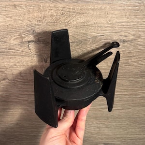 May include: A black, cast iron object with a circular centre and wing-like extensions. The object has a handle and is held in a hand. The object appears to be a vintage or antique piece.