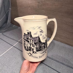 May include: A cream-colored ceramic pitcher featuring a black ink-style illustration of a building on Broadway High Street. The pitcher has a curved handle, a wide pouring spout, and a gold-toned rim. The design depicts a multi-story structure.