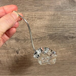 Puede incluir: Una cuchara de servir de plata con un diseño floral decorativo en el cuenco. El mango es curvo y la cuchara se sostiene en una mano.