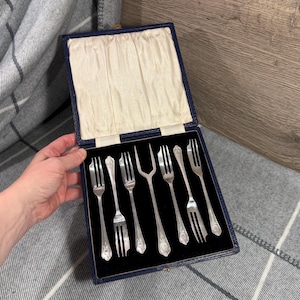 Op de afbeelding: Een set zilveren vorken in een blauwe doos met een crèmekleurige satijnen voering. De vorken hebben decoratieve bloempatronen op de handvatten. De doos is open en toont de opstelling van de vorken.