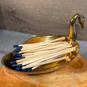 Op de afbeelding: Een gouden zwanen-vormige luciferdoos met een stapel houten lucifers met blauwe puntjes.