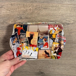Puede incluir: Una bandeja rectangular de metal con borde blanco y un collage de anuncios franceses vintage. La bandeja presenta imágenes de la Torre Eiffel, el Moulin Rouge y el Chat Noir, con texto en francés. La bandeja está sostenida por una mano.