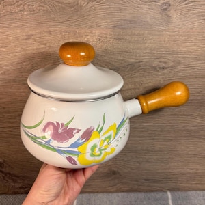 May include: White enamel saucepan with a floral design featuring yellow, pink, and green flowers. The saucepan has a wooden handle and lid.