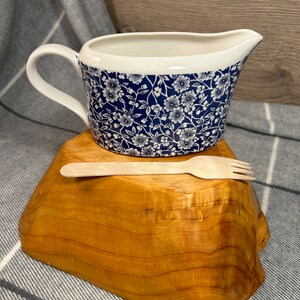 May include: A white ceramic gravy boat with a blue and white floral pattern. The boat is sitting on a wooden stand with a wooden spoon and fork.