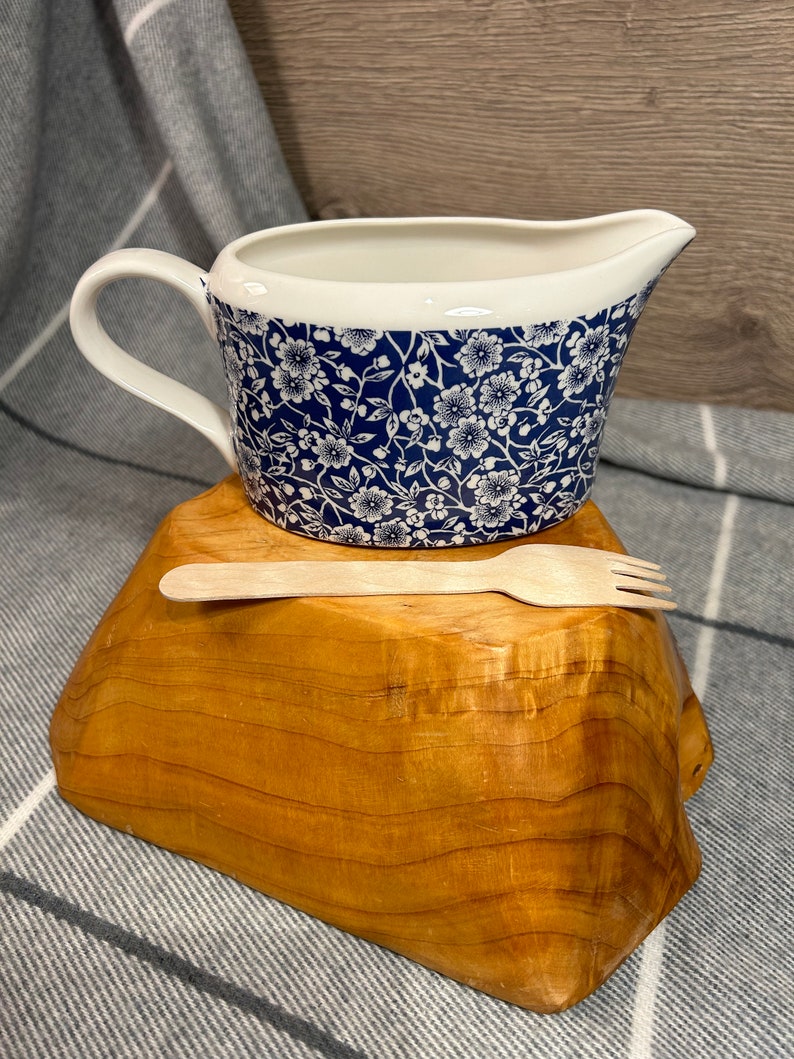 May include: A white ceramic gravy boat with a blue and white floral pattern. The boat is sitting on a wooden stand with a wooden spoon and fork.