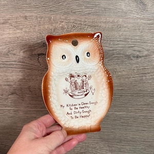 Puede incluir: Decoración de cocina de cerámica con forma de búho, con una combinación de colores marrón y blanco. El búho tiene ojos grandes y un pico pequeño. El texto "My Kitchen is Clean Enough To Be Healthy And Dirty Enough To Be Happy!" está impreso en la parte delantera.