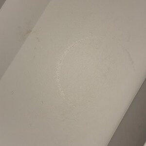 May include: A white, flat surface featuring a faint, circular ring stain in the center. The surface has a smooth, matte texture with subtle signs of wear, including minor smudges and light discoloration marks visible across the white area.