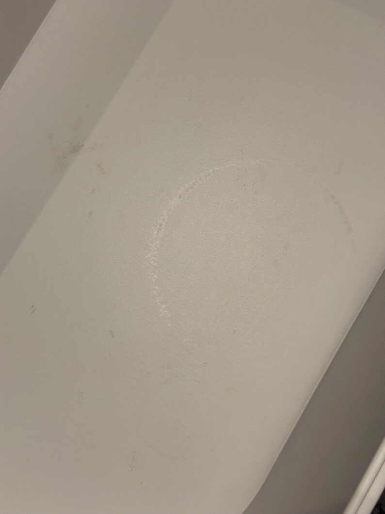 May include: A white, flat surface featuring a faint, circular ring stain in the center. The surface has a smooth, matte texture with subtle signs of wear, including minor smudges and light discoloration marks visible across the white area.