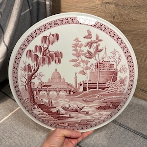 Peut inclure: Une grande assiette ronde en céramique blanche avec un motif rose et blanc. Le motif représente une scène avec un pont, un bâtiment avec un dôme et des arbres. L'assiette a une bordure décorative.