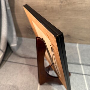 May include: A framed artwork with a black frame, displayed on a wooden easel. The artwork has a light-colored interior and is held in place with a wire. The easel is a dark brown color.