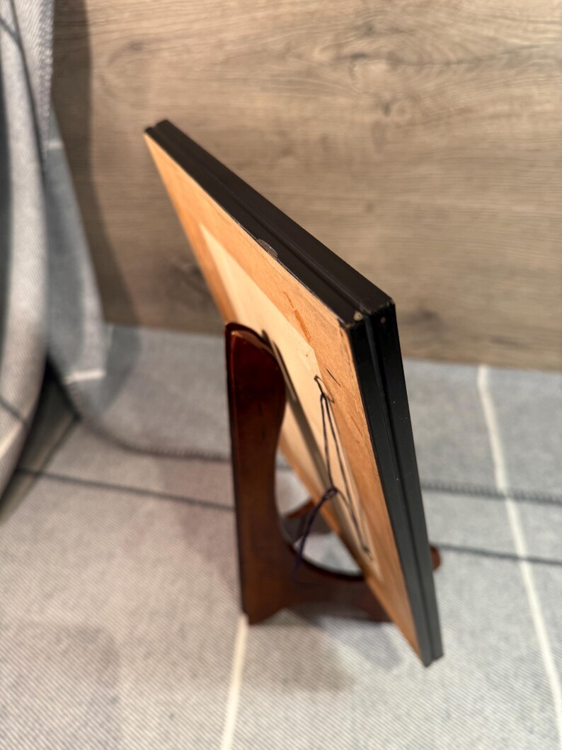 May include: A framed artwork with a black frame, displayed on a wooden easel. The artwork has a light-colored interior and is held in place with a wire. The easel is a dark brown color.