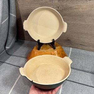 May include: Two off-white ceramic baking dishes with handles. One dish is on a black stand, and the other is below. The bottom dish has a dark green exterior. The dishes are round and appear to be vintage.