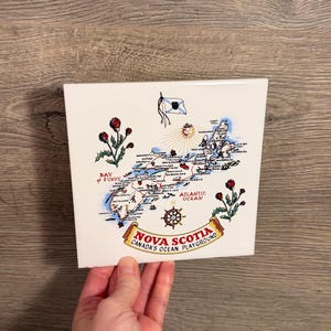 May include: A square white ceramic tile featuring a colourful illustrated map of Nova Scotia, Canada. The design includes red thistle flowers, a ship's wheel, a flag, and the text Nova Scotia Canada's Ocean Playground with Bay of Fundy labels.