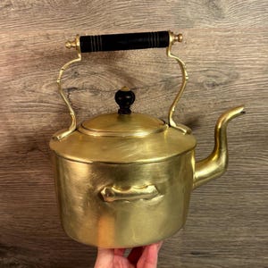 May include: A vintage brass kettle with a black wooden handle and lid knob. The kettle has a curved spout and decorative handle supports. The brass has a warm, golden hue, suggesting age and use. A classic kitchenware item.