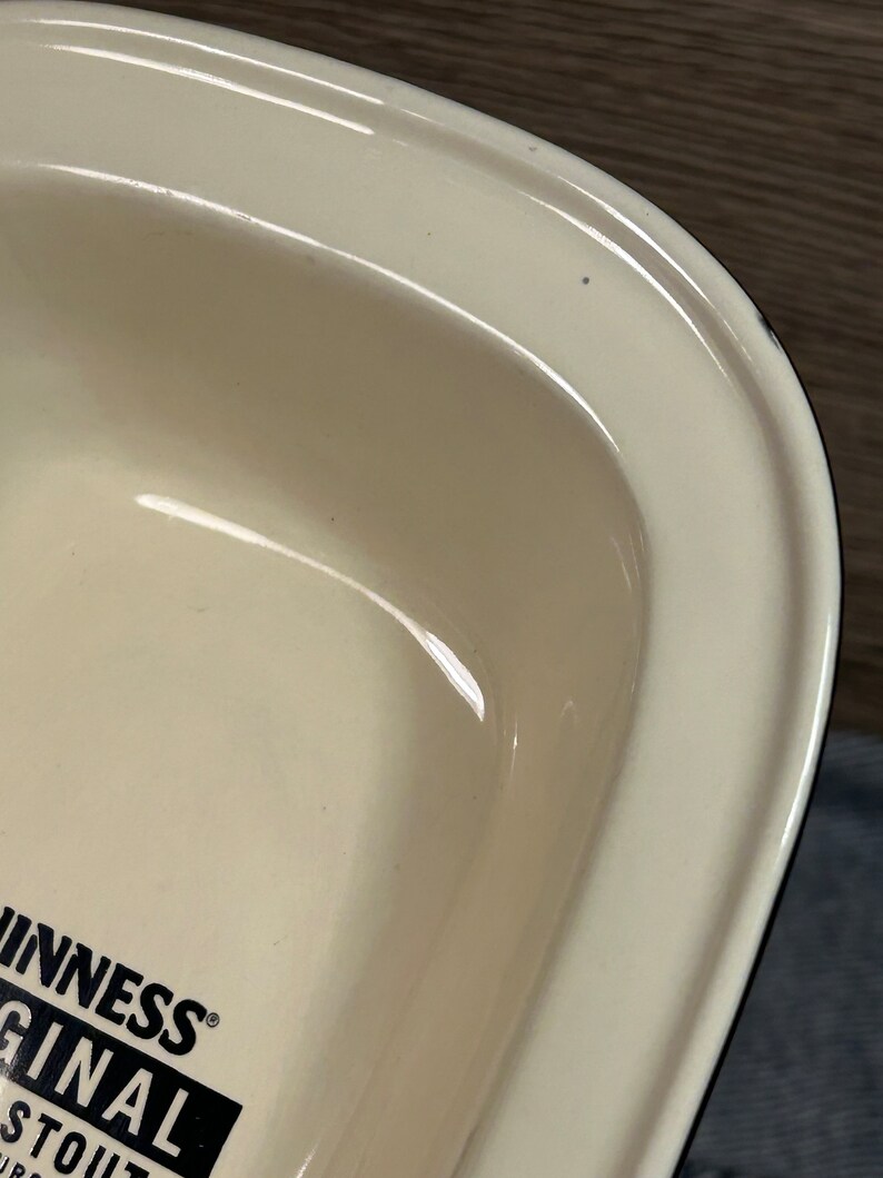 May include: Cream-colored, rectangular serving dish with rounded edges. The dish has a glossy finish and a black and white logo that reads "Guinness Original Stout". The dish is likely made of ceramic or porcelain.