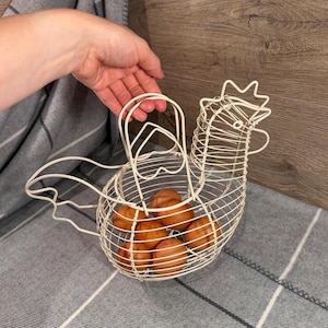 Op de afbeelding: Een crèmekleurige draadmand in de vorm van een kip met een handvat aan de bovenkant. De mand bevat zes bruine eieren. De draadstructuur vormt de omtrek van een kip met een kam, snavel en staartveren, wat een open opbergmand vormt.