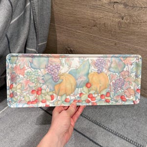 Könnte beinhalten: Rechteckige Serviertablett mit farbenfrohem botanischem Design. Das Tablett zeigt Illustrationen von Kürbissen, Trauben und Beeren in Orange, Lila und Rot, vor dem Hintergrund grüner und pastellfarbener Blätter.