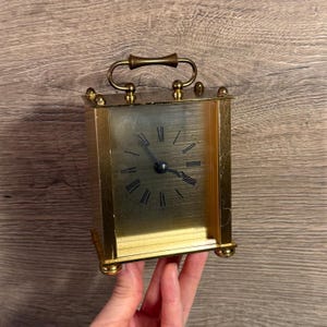 May include: A gold-coloured rectangular travel clock with a handle and Roman numeral hour markers. The clock has a gold frame and a black clock face. The clock is resting on a wooden surface.