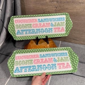 May include: Two rectangular serving trays with a white center and green polka dot borders. The trays have the words "CUCUMBER SANDWICHES, SCONE CREAM & JAM, AFTERNOON TEA" in colorful lettering. The trays are on a wooden surface.