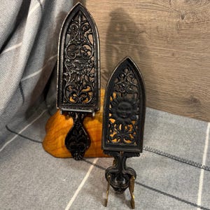 May include: Two black cast iron trivets with intricate designs. The larger trivet features a sun face with a floral design. The smaller trivet features a floral design with a face in the center.