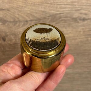 May include: A small, round, gold-colored trinket box with a white ceramic lid featuring a black and brown illustration of a tree and a fence.