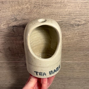 May include: A beige ceramic tea bag holder with a rounded shape and a small hole at the top. The words "TEA BAGS" are written in blue on the side. The holder is held by a hand.