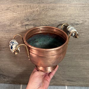 Può includere: Una ciotola di rame con due manici. I manici sono in ottone e hanno impugnature in ceramica bianca con disegni floreali blu. La ciotola ha una base rotonda e un bordo leggermente svasato. L'interno della ciotola ha una patina verde.